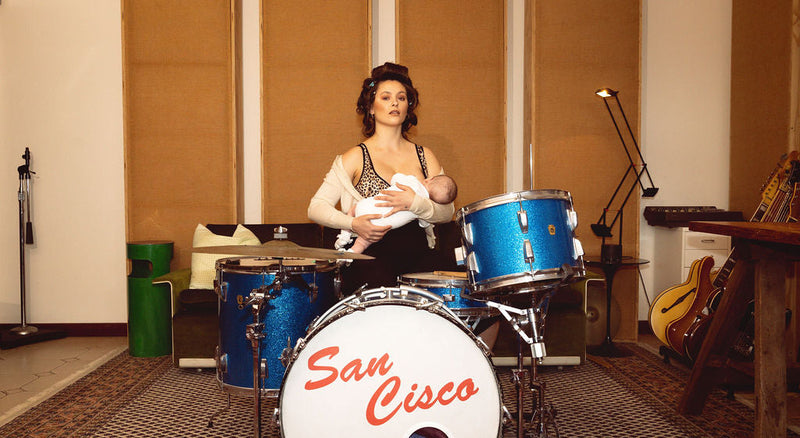 Woman holding a baby in front of a drum set with 'San Cisco' branding in a studio setting.