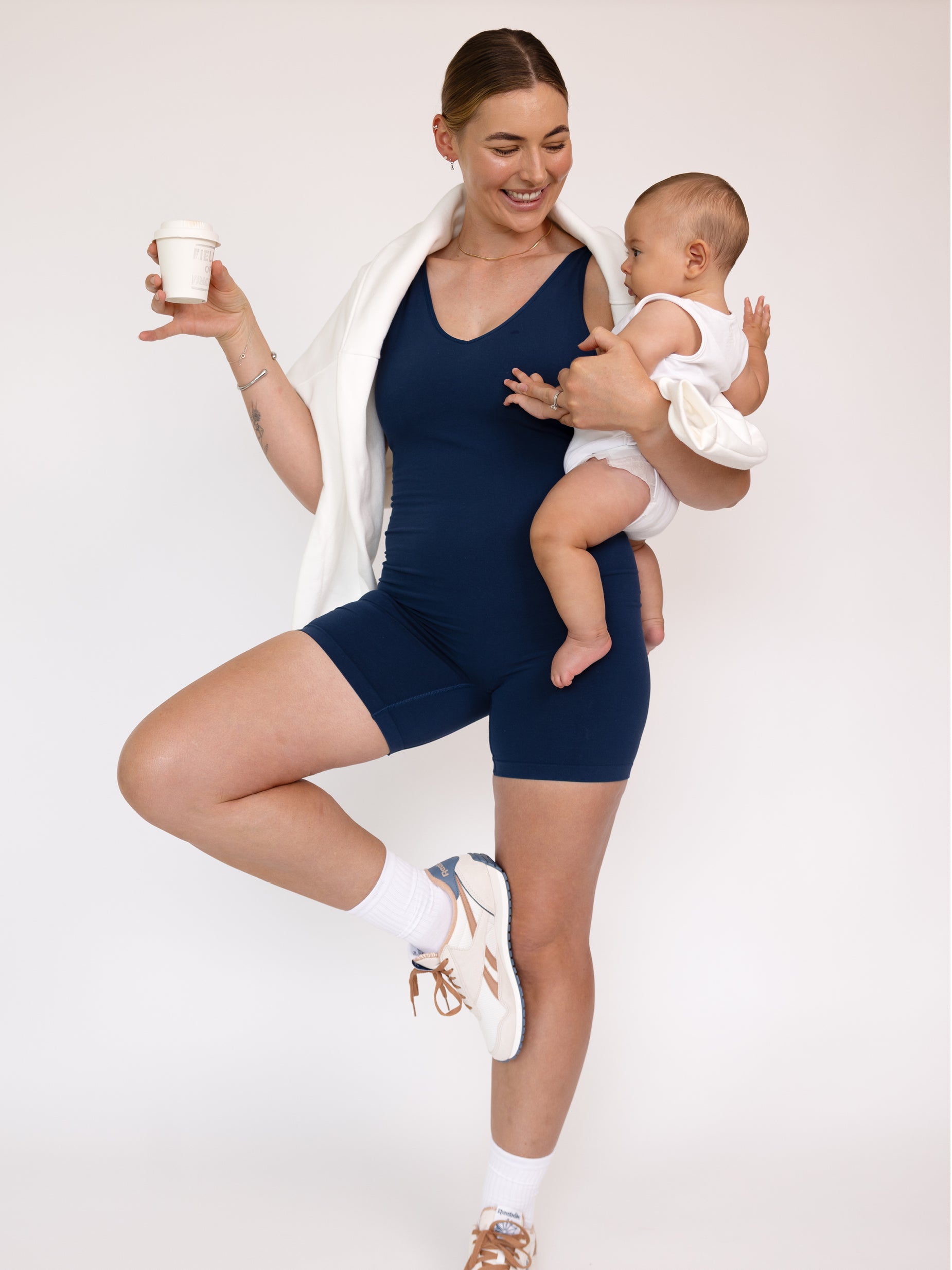 Woman in navy athletic outfit holding a baby and a cup against a white background
