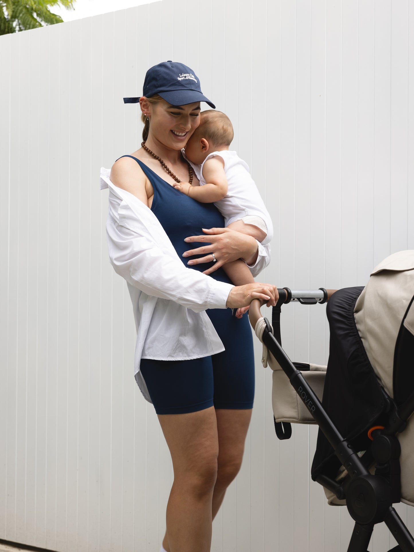 Woman in athletic wear holding a baby next to a stroller against a white wall.