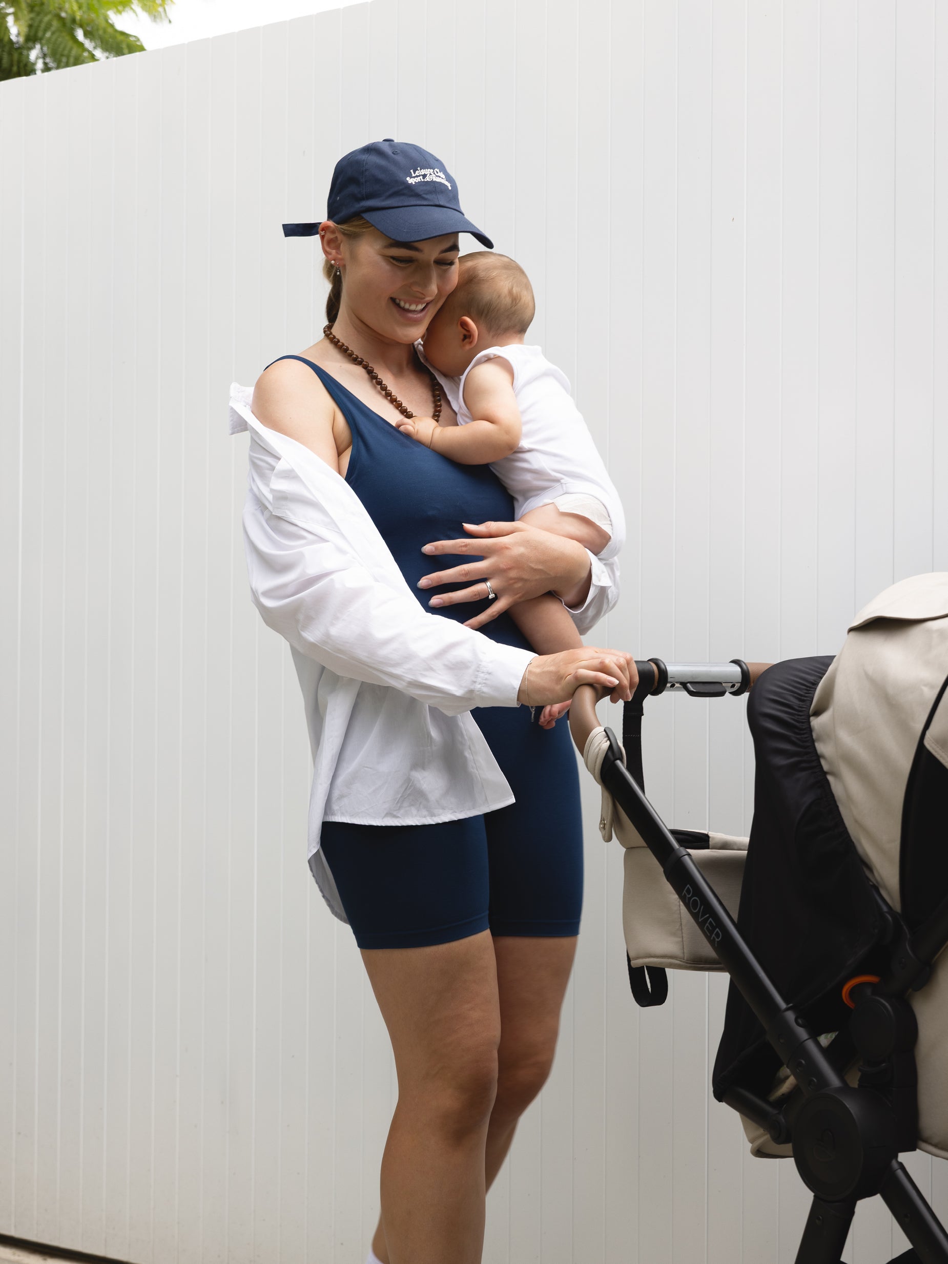Woman in athletic wear holding a baby next to a stroller against a white wall.