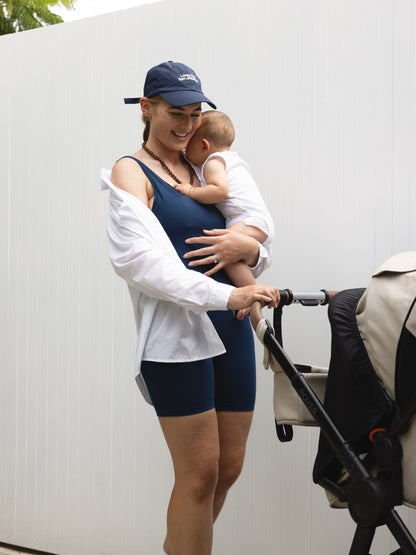 Woman in athletic wear holding a baby next to a stroller against a white wall.