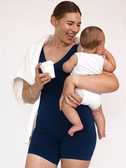 Woman in a navy blue bodysuit holding a baby and a white cup against a plain background